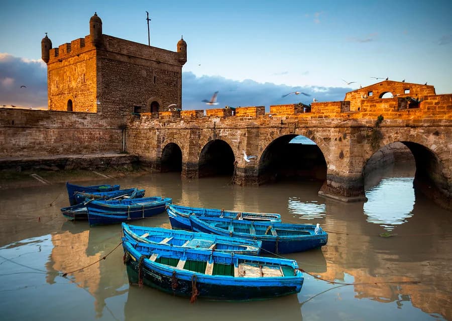 Blue fishing boats moored beneath the stone ramparts of the Skala du Port in Essaouira at dusk